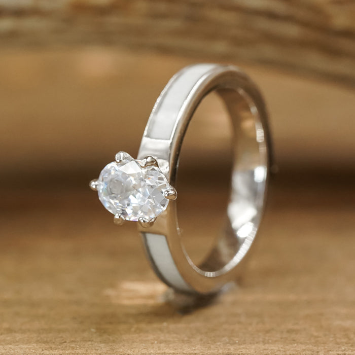 A sterling silver engagement ring named The Grand Adair, featuring a white antler inlay and a single round-cut clear gemstone set in a prong setting, rests gracefully on a wooden surface with a blurred background.
