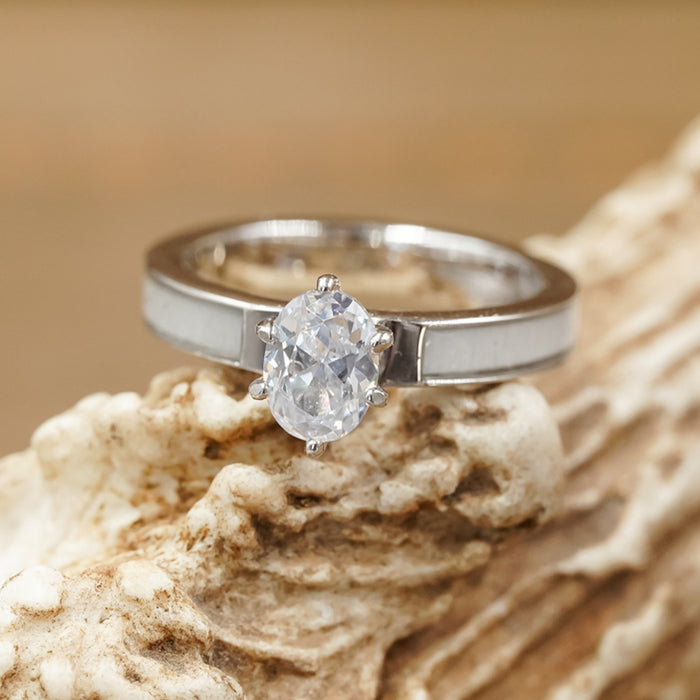 The Grand Adair - White Antler Inlay engagement ring, featuring a sterling silver band with an oval cut cubic zirconia, rests on a textured, light-colored surface reminiscent of coral or rock. The softly blurred background glows in warm tones.