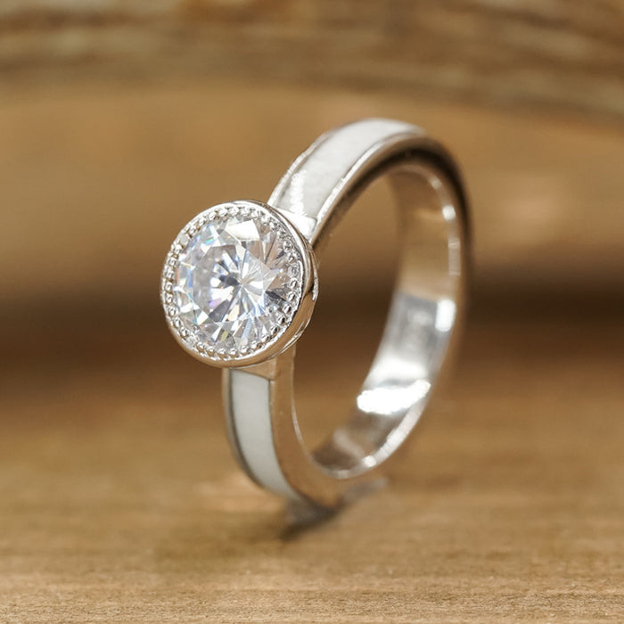The Grand Avonelle - White Antler Inlay is a sterling silver ring boasting a round, clear cubic zirconia gemstone in a bezel setting, accented by a striking white antler inlay on the band and elegantly displayed on a wooden surface.