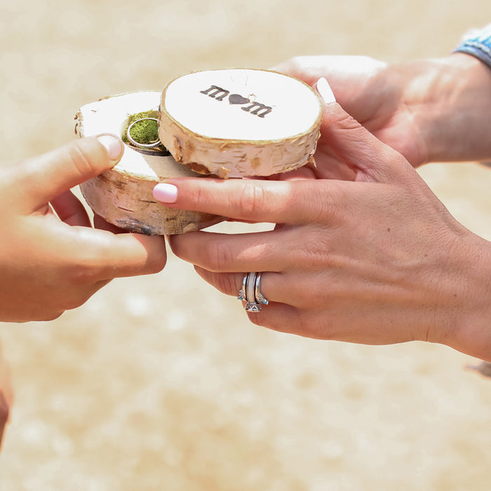 Two people hold a small wooden ring box with "mom" and a heart engraved on the lid. One hand opens the box, revealing The Grand Avonelle - White Antler Inlay engagement ring with a stunning green cubic zirconia stone inside. The background is softly blurred.