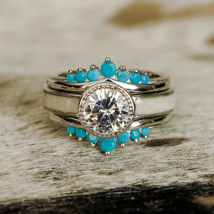 The Grand Avonelle - White Antler Inlay is a sterling silver ring featuring a large central clear cubic zirconia gemstone, framed by small turquoise stones arranged in a semicircle around the base, beautifully displayed on a textured surface.