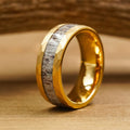 The Great Sand Dunes is a gold ring featuring a textured, faceted surface and a central inlay of grayish, marbled antler, shown upright on a wooden surface with a softly blurred wooden background.