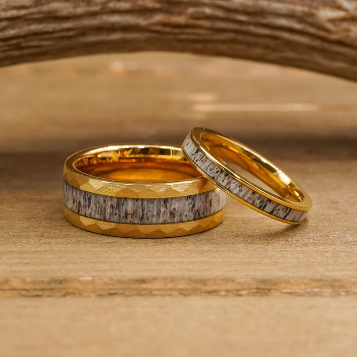 Two gold rings from "The Great Sand Dunes" collection, featuring a gray, wood-textured inlay, rest on a wooden surface. The larger ring lies flat while the smaller one leans beside it, highlighting the distinctive character of unique antler jewelry.
