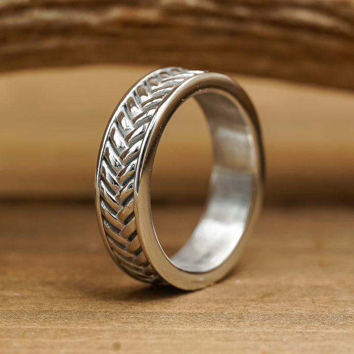 The Herringbone, a silver stacking band featuring a distinctive engraved herringbone pattern encircling the band, is displayed upright on a wooden surface against a softly blurred wooden background.