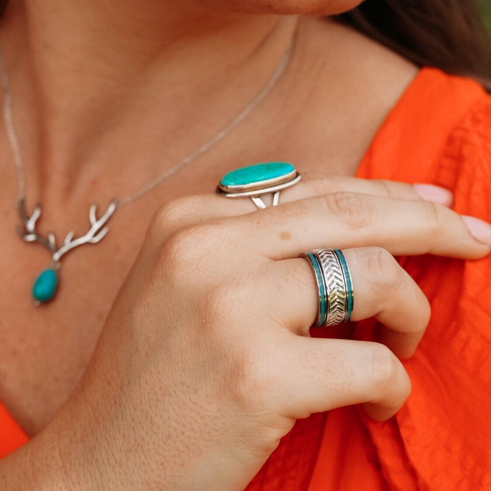 A woman wearing an orange top shows her hand adorned with turquoise rings and a sleek silver ring. She also wears The Herringbone necklace with a turquoise pendant, completing the look with a touch of elegance.