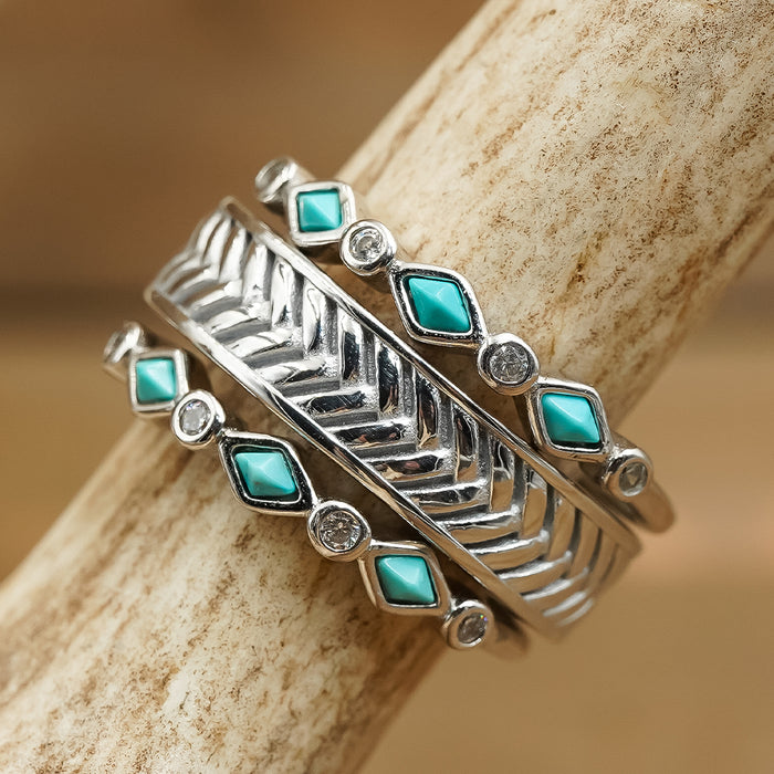 Three silver rings, including The Herringbone stacking band and two eye-catching bands with geometric patterns—each adorned with turquoise and clear gemstones—stacked together on a textured, light tan surface.