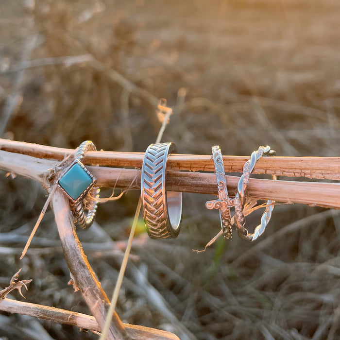 Four silver rings, including The Herringbone band and a turquoise ring, rest on a dry plant stem outdoors in warm sunlight, with blurred grass in the background.