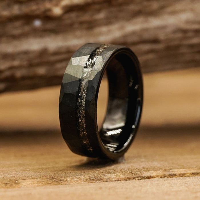 The Hoba, a black faceted tungsten carbide ring featuring a textured, glittery stripe encircling its center, stands upright on a wooden surface with a blurred wood background.