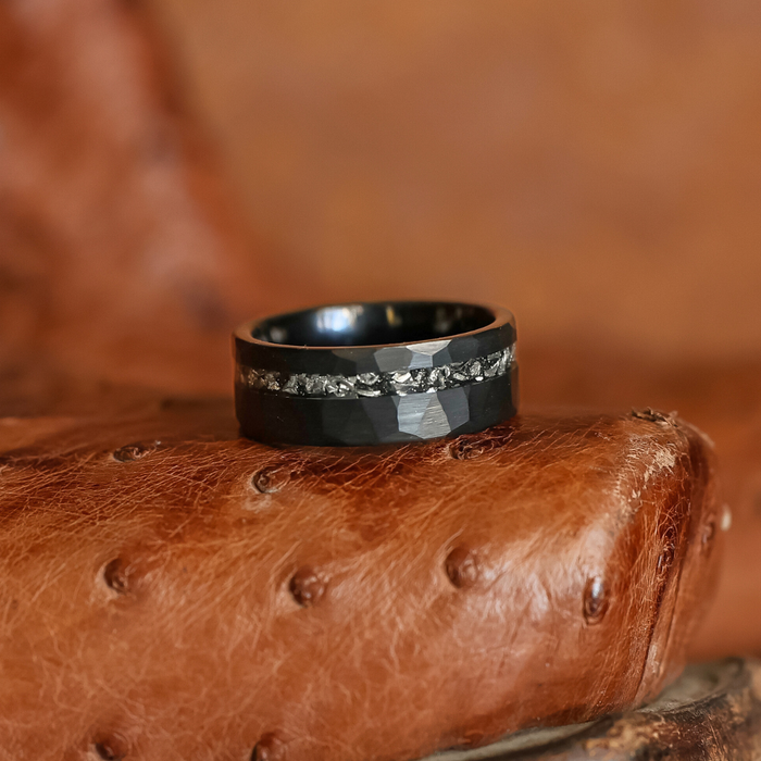 The Hoba, a black tungsten carbide ring with a textured, diamond-like band, rests on brown, textured leather upholstery.