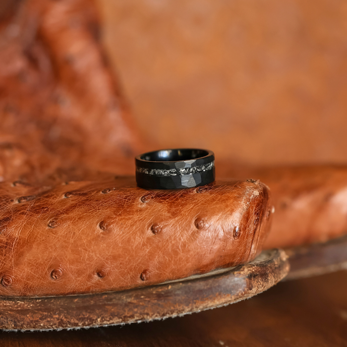 The Hoba, a black tungsten carbide ring featuring a hammered finish and textured center band, rests on the arm of a worn brown leather chair with a distinctive ostrich pattern.