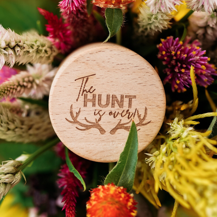 The Hunt Is Over" Ring Box—a wooden, round box engraved with "The Hunt Is Over" and decorated with antlers—rests amid vibrant flowers and greenery, making it a charming keepsake for an Antler Ring presentation or a unique addition to your collection.
