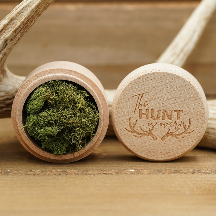 The Hunt Is Over" Ring Box: A small, round beech wood box with a lid engraved "The Hunt is Over" and antlers. The box is open, displaying green moss inside—ideal for presenting an antler ring. Both the box and the ring are set on a wooden surface, accented by deer antlers in the background.