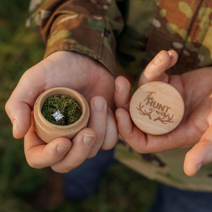 A person in camouflage holds "The Hunt Is Over" Ring Box, crafted from beech wood and lined with moss, featuring an antler ring inside. The lid displays the phrase "The HUNT is over" accented by elegant antler designs.