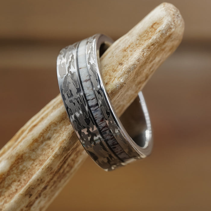 The Kings Canyon ring, featuring a silver antler design with a camouflage pattern and a light inlay band, is showcased on a textured, bone-like surface against a softly blurred brown background. Perfect as a men's wedding band, this distinctive piece blends rugged appeal with contemporary style.