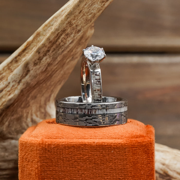 Two silver rings, including The Kings Canyon—a striking men’s wedding band with engraved patterns—are displayed upright on an orange velvet box. One ring features a round diamond, while textured wood in the background adds warmth to the scene.