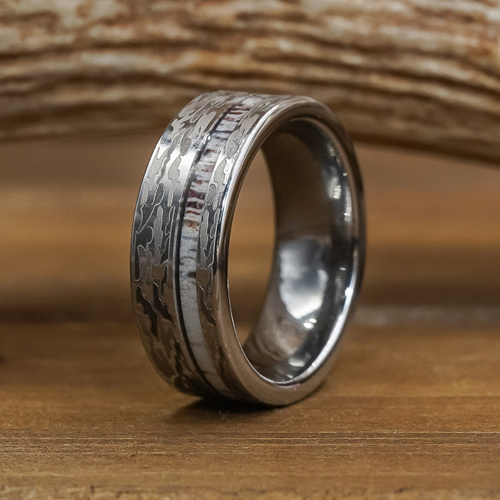 A men's wedding band called "The Kings Canyon," featuring a silver, hammered metal design with a wood-like textured background, is displayed upright on a wooden surface.