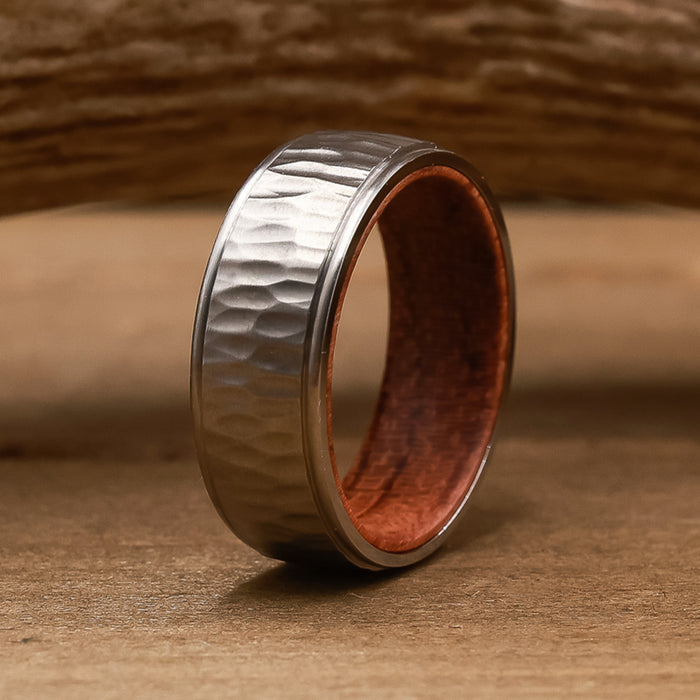 Introducing The Koa: a silver hammered metal ring featuring a textured surface and a polished reddish-brown inner band. Standing upright on a wooden surface with a blurred wood background, The Koa showcases striking contrast and refined craftsmanship, making it the ideal men's wedding band for those seeking distinctive style.