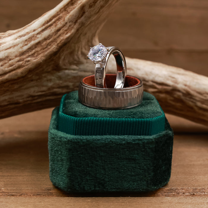 Two rings—a diamond engagement ring and a textured wedding band—are displayed on a green velvet box, with The Koa, a unique men's wedding band featuring koa wood and titanium, nestled beside them. A wooden antler prop adds rustic charm to the background.