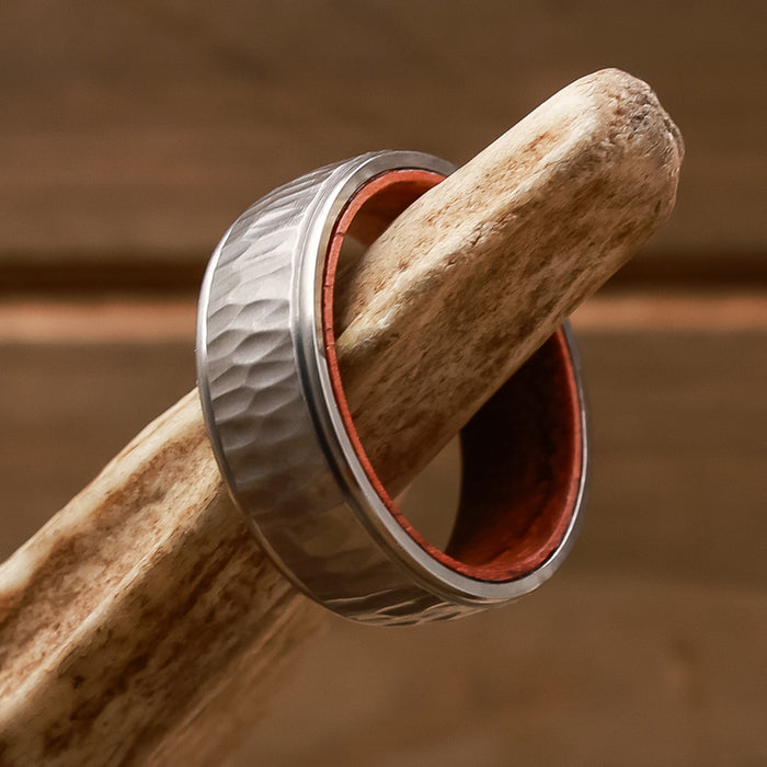 The Koa, a silver hammered metal ring featuring a striking red inner lining and reminiscent of a unique men's wedding band, is displayed atop a piece of antler set against a wooden background.