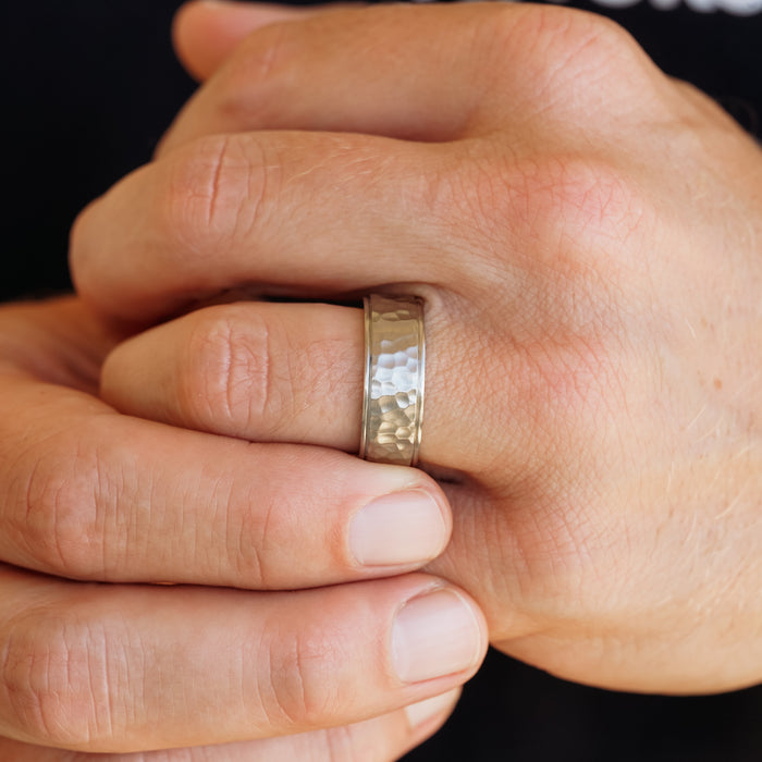 A close-up of a person’s hand wearing The Larry ring—a distinctive hammered metal band—on their finger, while the other hand gently holds the finger. The background is softly out of focus.