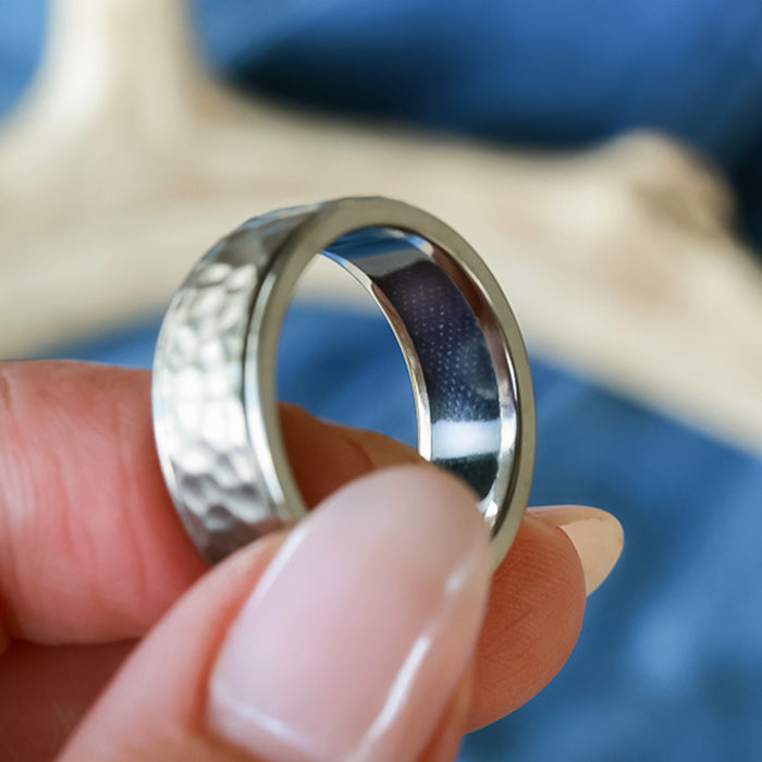 A hand with manicured nails holds The Larry, a silver hammered metal titanium ring, against a blurred blue and beige background.