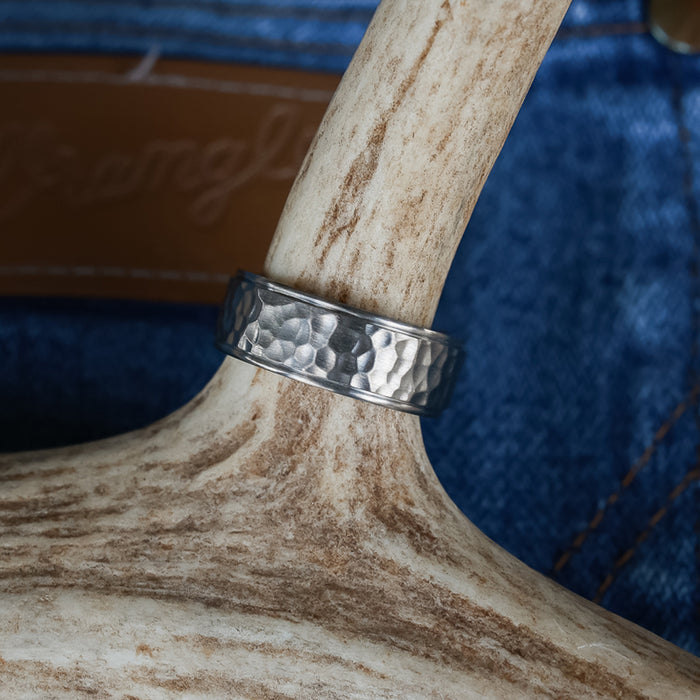 A silver hammered titanium ring, named The Larry, rests on a light antler, with blue denim fabric and a brown leather patch softly blurred in the background—blending rugged style with unique inspiration from antler rings.