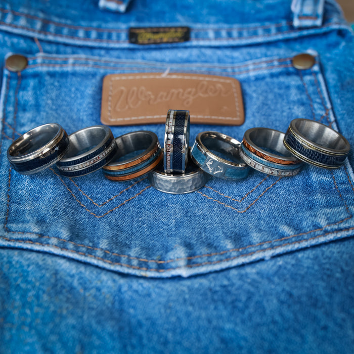 Seven rings with various designs—including The Larry, an elegant antler ring, and a sleek titanium ring—rest on the back pocket of blue Wrangler jeans, with the leather brand label visible above the pocket.