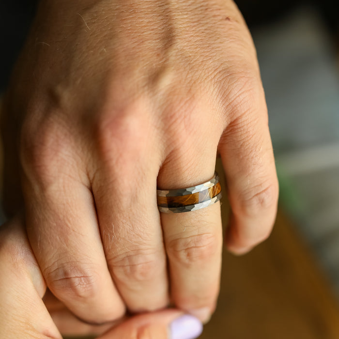 A close-up showcases a hand adorned with The Lawrenceburg, a faceted tungsten carbide ring featuring shades of brown, gray, and silver that evoke subtle elegance. Another hand gently holds one of the fingers, set against a softly blurred background.