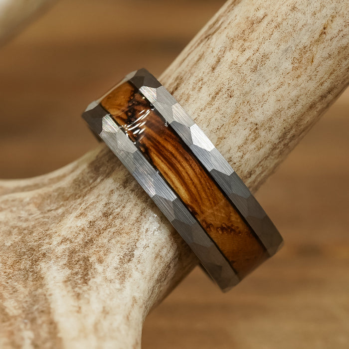 Introducing The Lawrenceburg: a faceted tungsten carbide ring featuring a polished wood inlay reminiscent of a whiskey barrel, beautifully displayed on a light-colored antler with a blurred brown background.