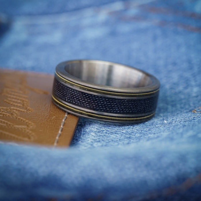 The LeDoux, a silver and dark blue tungsten carbide ring with metallic edges, rests on blue denim fabric beside a piece of brown leather.