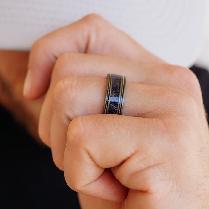 A close-up of a hand wearing The LeDoux, a black and silver tungsten carbide ring, on the middle finger while holding a white object near the face.