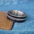 Introducing The Leon: a stylish men’s band, this silver ring featuring black and white inlay is displayed on a tan leather patch against classic blue denim fabric.