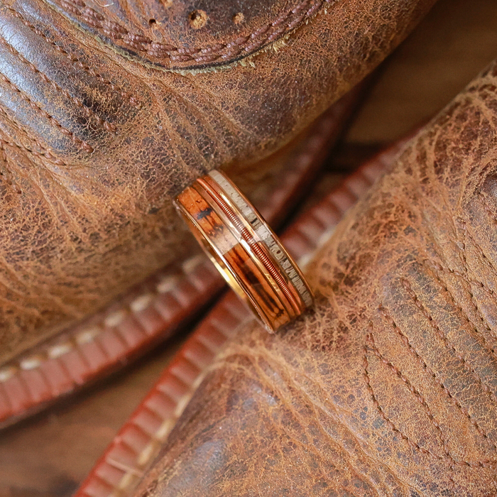 The Letchworth, a rose gold ring with metal accents, rests on a pair of worn, brown cowboy boots featuring visible stitching and textured leather.