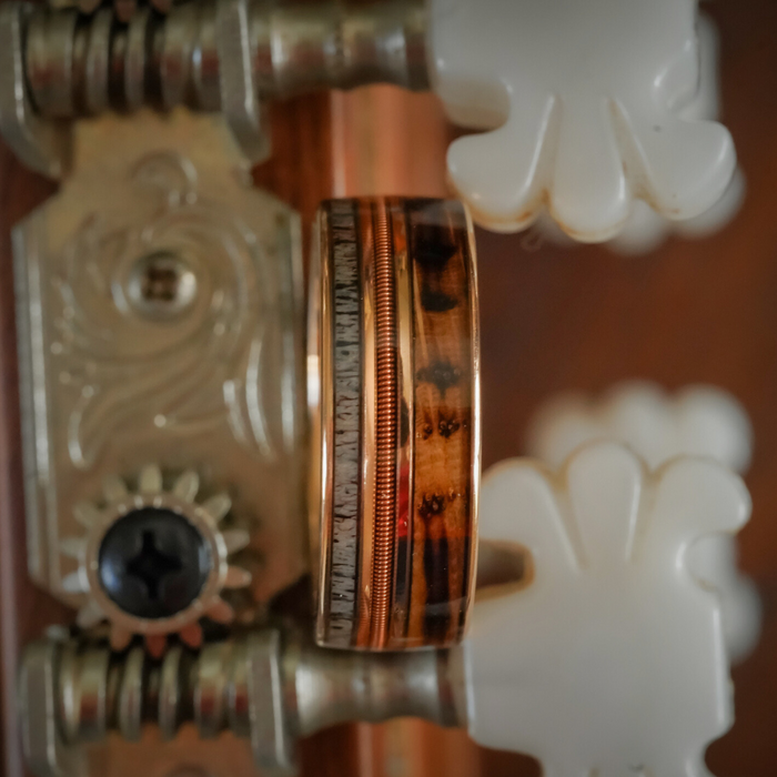 The Letchworth, a tungsten carbide ring featuring a wooden inlay and metallic edging, rests on the tuning peg of a stringed instrument—possibly a guitar—with ornate metal and white plastic details in view.