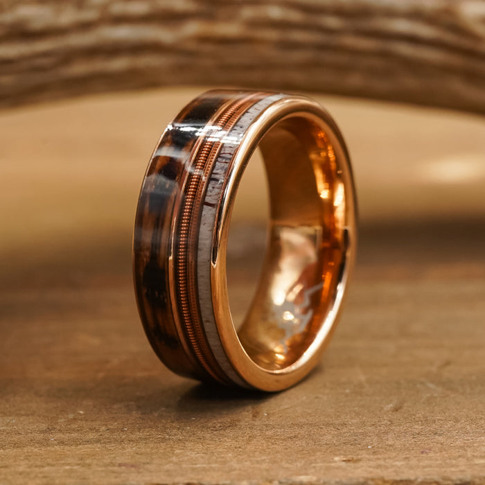 The Letchworth, a tungsten carbide ring with dark brown and silver inlays featuring intricate detailing, is displayed upright on a wooden surface with a rustic wood background.