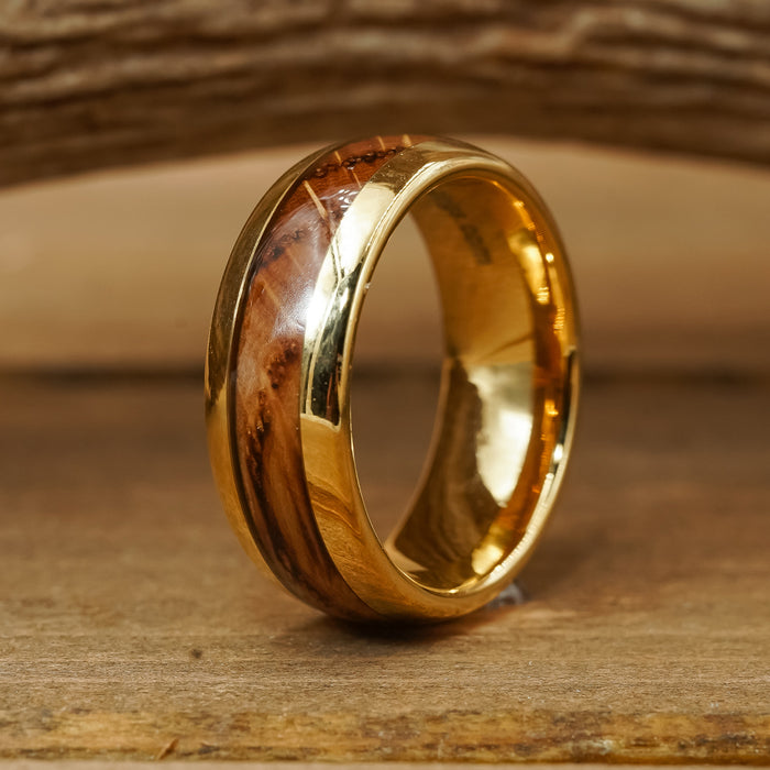 The Loretto, a gold tungsten ring featuring a polished wood inlay, stands upright on a wooden surface against a softly blurred wooden background, making it an elegant choice for a men's wedding band.