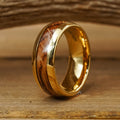 The Loretto, a gold tungsten ring featuring a polished wood inlay, stands upright on a wooden surface against a softly blurred wooden background, making it an elegant choice for a men's wedding band.