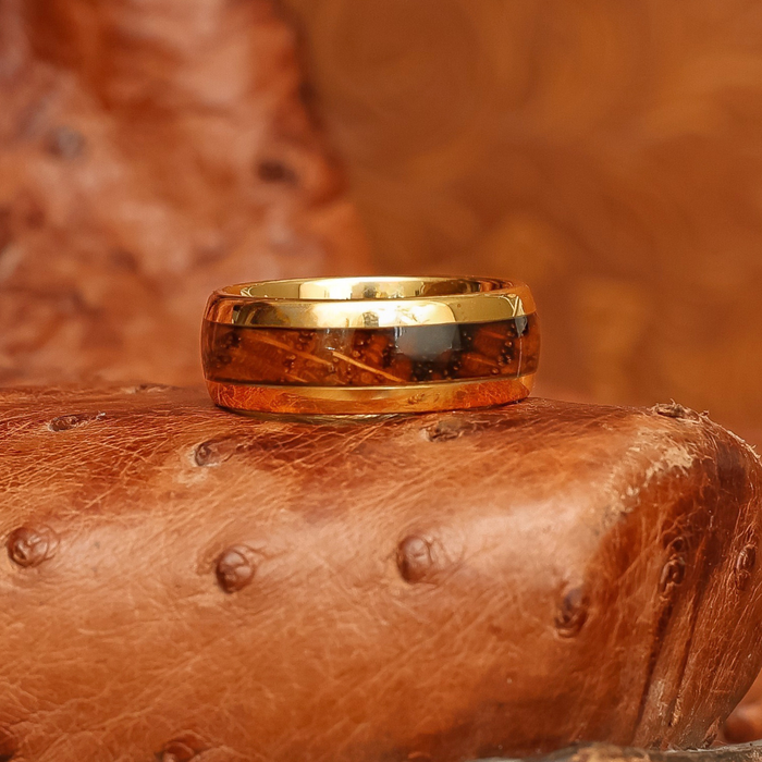 The Loretto—a gold tungsten ring featuring a brown wood inlay that evokes the look of a whiskey barrel—rests atop a dimpled, textured brown leather surface.