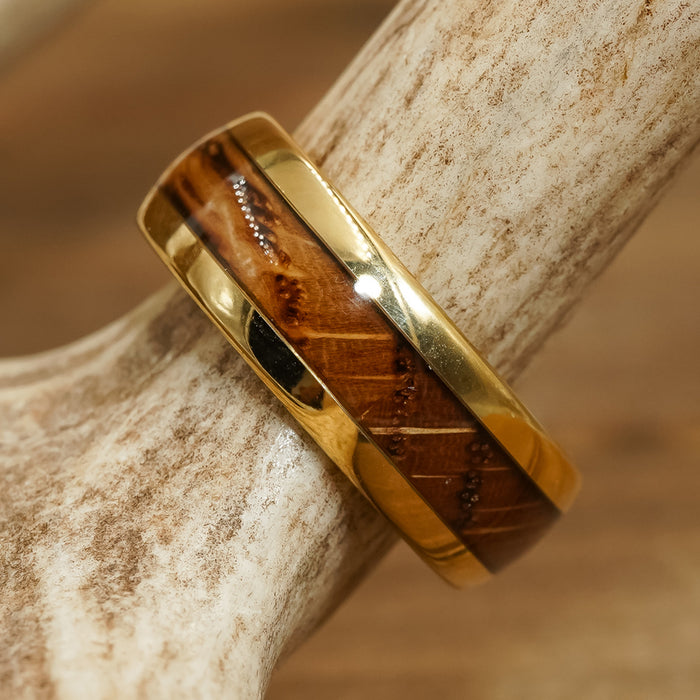 The Loretto, a gold tungsten ring featuring a polished wooden inlay inspired by classic whiskey barrel rings, rests on a textured, light-colored antler. The background is softly blurred in warm brown tones.