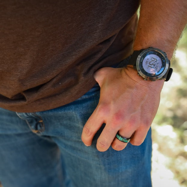 A person wearing a brown shirt, blue jeans, a black sports watch, and The Los Glaciares—a unique men's ring with turquoise inlay—stands outdoors with their right hand resting near their pocket.