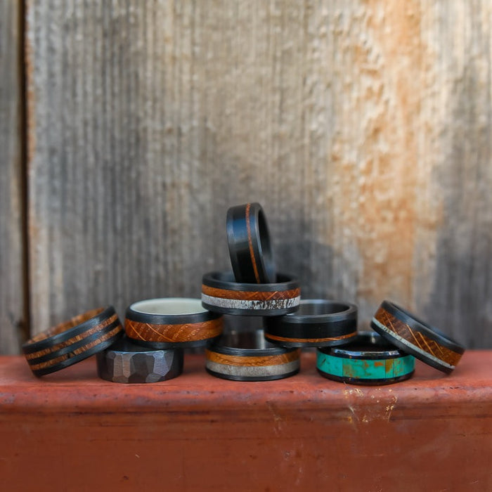 A group of eleven unique rings, including The Los Glaciares—a striking turquoise inlay ring—and a bold black ceramic ring, is arranged on a ledge against a rustic wooden background. The collection showcases distinctive wood, stone, and metal designs, making it an ideal selection for anyone seeking a unique men’s ring.