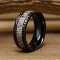 The Lowell is a men's hammered ring with a black finish, featuring a light-colored, antler-inspired wood-like inlay. It is displayed on a wooden surface with a softly blurred wooden background.
