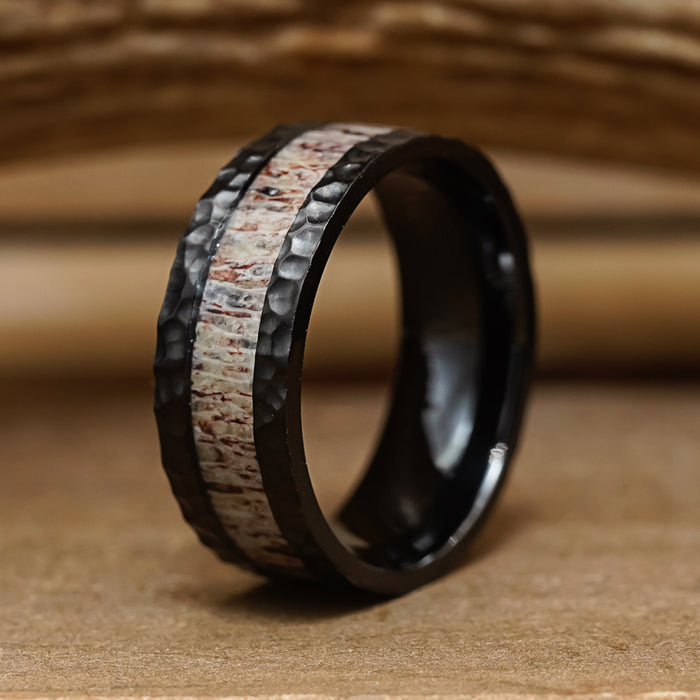 The Lowell is a men's hammered ring with a black finish, featuring a light-colored, antler-inspired wood-like inlay. It is displayed on a wooden surface with a softly blurred wooden background.