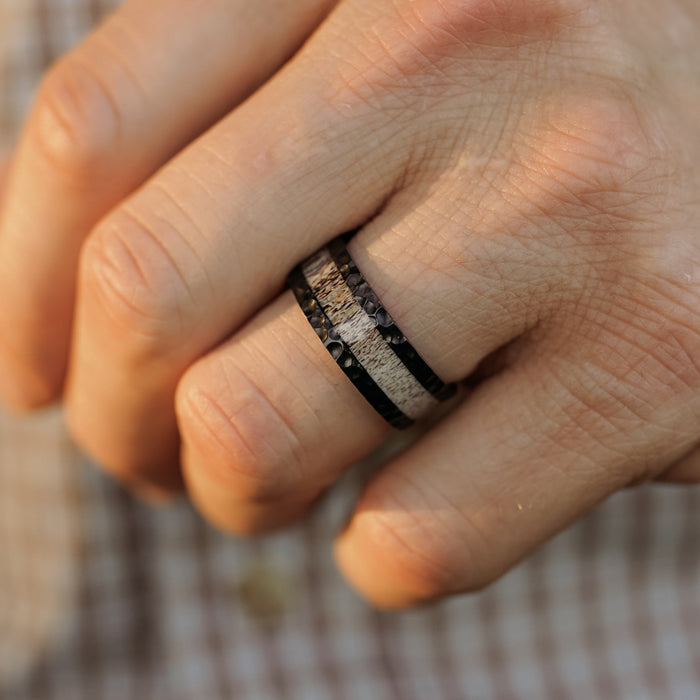 A close-up of a hand wearing The Lowell, a black men's hammered ring featuring silver and textured inlay details, with a plaid shirt visible in the background.