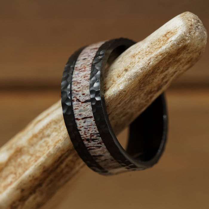 The Lowell, a black men’s hammered ring featuring a marbled, reddish-white inlay reminiscent of antler grain, is displayed on a piece of bone against a brown background.