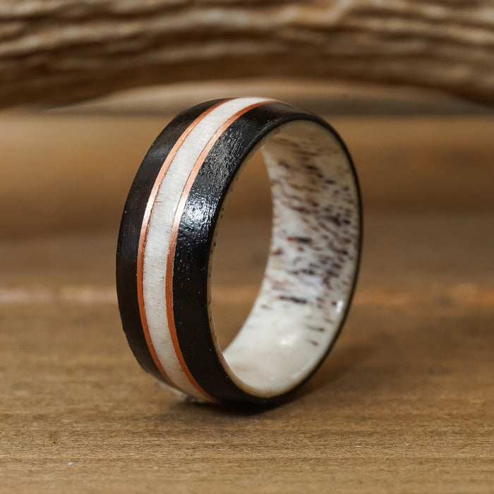 The Loxahatchee, a rustic wedding band with a black outer edge, a copper stripe, and a speckled white central antler inlay, is displayed upright on a wooden surface.