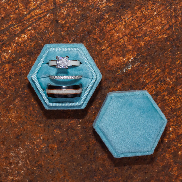 The Loxahatchee, a set of three rings featuring two sleek silver bands and one rustic wedding band adorned with a square-cut diamond, is elegantly presented in a hexagonal light blue velvet box, with the lid resting nearby on a textured rusty brown surface.
