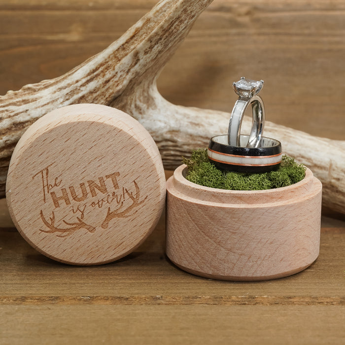 Two rings, including The Loxahatchee rustic wedding band, rest on green moss inside an open wooden ring box. The box lid, set beside it, is engraved with the words "The Hunt is Over" alongside antler designs, while a deer antler accents the background.