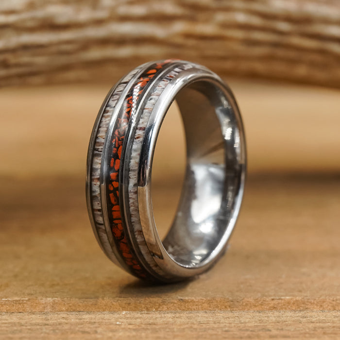 Introducing The Makoshika: a silver tungsten carbide ring featuring two inlaid bands—a textured, light wood-toned band and a slim, dark band accented with orange flecks reminiscent of red dinosaur bone—displayed on a wooden surface against a softly blurred background.