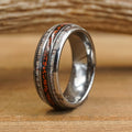 Introducing The Makoshika: a silver tungsten carbide ring featuring two inlaid bands—a textured, light wood-toned band and a slim, dark band accented with orange flecks reminiscent of red dinosaur bone—displayed on a wooden surface against a softly blurred background.